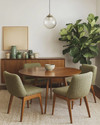 A cozy Mid-Century Modern dining room featuring a round walnut dining table with four upholstered chairs in a muted olive green boucle fabric. A George Nelson bubble lamp hangs above, casting a soft glow. A vintage teak credenza against the wall displays ceramic vases and a large Fiddle Leaf Fig plant. — styling and décor detail