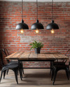 Industrial breakfast nook featuring a reclaimed wood table, black metal chairs, and three matte black dome pendant lights hanging low. An exposed red brick wall provides a textured backdrop, and an exposed Edison bulb sconce adds warm, ambient lighting. — styling and décor detail