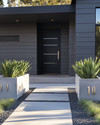 A modern exterior featuring a fresh coat of deep charcoal grey paint on the siding, a contrasting matte black front door, and sleek brushed nickel house numbers. The pathway combines light grey concrete pavers with dark pea gravel, and large concrete planters flank the entrance with sculptural green plants. — styling and décor detail