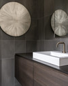 A modern powder room features a floating dark wood vanity with an integrated white quartz sink and brushed nickel faucet, set against large format charcoal grey porcelain tiles. A simple round mirror and abstract metal wall art complete the sleek, resilient design. — alternate angle