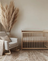 A serene Japandi baby room with a light wood crib, a soft grey glider, and a neutral rug, accented by a tall dried pampas grass arrangement in a ceramic vase and a subtly patterned throw blanket. — close-up of textures and materials