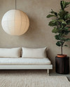 A serene Japandi poolhouse interior featuring a low-slung white oak daybed with natural linen cushions, a dark wenge wood side table, and a large, round paper lantern pendant light. The walls are a soft greige, complemented by a subtle textured rug in cream and a tall, slender fiddle leaf fig plant in a terracotta pot. — close-up of textures and materials