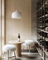 A small Scandinavian wine cellar with light ash wood shelving, a light gray concrete floor, and matte black metal wine racks. A minimalist white pendant light hangs over a small, round light wood tasting table with two simple stools covered in white sheepskin throws. — styling and décor detail