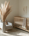 A serene Japandi baby room with a light wood crib, a soft grey glider, and a neutral rug, accented by a tall dried pampas grass arrangement in a ceramic vase and a subtly patterned throw blanket.