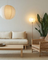 A serene Japandi basement living area with a low-profile cream sofa, a light wood coffee table, and a woven rattan armchair. Warm diffused lighting comes from a paper lantern pendant and a minimalist floor lamp, while a large snake plant in a ceramic pot adds natural greenery. — alternate angle