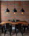 Industrial breakfast nook featuring a reclaimed wood table, black metal chairs, and three matte black dome pendant lights hanging low. An exposed red brick wall provides a textured backdrop, and an exposed Edison bulb sconce adds warm, ambient lighting.