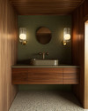 A Mid-Century Modern powder room featuring a floating walnut vanity, a small-chip terrazzo floor, and a muted olive green textured wallpaper, illuminated by brass and frosted glass sconces. — alternate angle