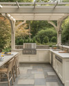 A refined traditional outdoor kitchen featuring off-white Shaker-style cabinetry, honed Pietra Cardosa countertops, and integrated stainless steel appliances under a simple pergola, with natural stone pavers. — styling and décor detail