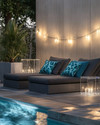 Modern poolside area with low-profile charcoal grey modular loungers, clear acrylic side tables, and large faux concrete planters. The scene is illuminated by sleek rechargeable LED lanterns and subtle solar-powered string lights, with deep teal and slate blue geometric outdoor cushions adding pops of color. — styling and décor detail