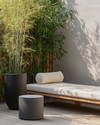 A small Japandi backyard featuring a low-profile teak bench with off-white cushions, a round grey stone side table, and a tall bamboo plant in a black ceramic planter, all set on light wood decking. — styling and décor detail