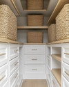 A clean coastal minimalist closet featuring light oak shelving, white built-in drawers with brushed nickel pulls, and tightly woven seagrass baskets, all bathed in natural light.