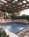 A cozy farmhouse pool area featuring a large swimming pool with natural stone coping, surrounded by reclaimed wood pergola. Plush outdoor sofas with cream cushions and striped pillows are arranged on a flagstone patio, illuminated by Edison bulb string lights overhead and decorative lanterns. — alternate angle