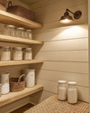 A warm farmhouse pantry with cream shiplap walls, natural pine shelves, and a semi-flush mount aged bronze light fixture illuminating clear glass jars and white ceramic canisters. A woven jute runner is on the floor. — styling and décor detail