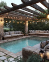 A cozy farmhouse pool area featuring a large swimming pool with natural stone coping, surrounded by reclaimed wood pergola. Plush outdoor sofas with cream cushions and striped pillows are arranged on a flagstone patio, illuminated by Edison bulb string lights overhead and decorative lanterns. — close-up of textures and materials