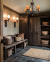 A rustic mudroom featuring warm layered lighting from a wrought iron chandelier and wall sconces, with slate floor tiles, a reclaimed wood bench, and dark wood storage cabinets, accented by woven baskets and a jute rug. — alternate angle