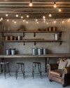 A rustic garage interior featuring exposed concrete walls, a salvaged wood workbench with metal stools, overhead string lights with Edison bulbs, and open shelving made from iron pipes and rough-sawn lumber, accented with galvanized buckets and a worn leather armchair.