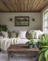 A rustic sunroom featuring a natural wood ceiling, a comfortable sofa with cream linen cushions, a chunky knit throw, and sage green accent pillows, surrounded by potted ferns and a vintage distressed wooden coffee table. — alternate angle