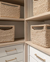 A clean coastal minimalist closet featuring light oak shelving, white built-in drawers with brushed nickel pulls, and tightly woven seagrass baskets, all bathed in natural light. — styling and décor detail