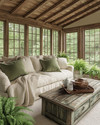 A rustic sunroom featuring a natural wood ceiling, a comfortable sofa with cream linen cushions, a chunky knit throw, and sage green accent pillows, surrounded by potted ferns and a vintage distressed wooden coffee table.