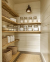 A warm farmhouse pantry with cream shiplap walls, natural pine shelves, and a semi-flush mount aged bronze light fixture illuminating clear glass jars and white ceramic canisters. A woven jute runner is on the floor.