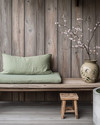 A Japandi porch blending vintage and modern, featuring a low-slung teak bench with a sage green linen cushion, a small hand-carved wooden stool as a side table, and a vintage Japanese stoneware vase with a single cherry blossom branch, all set against a backdrop of weathered wood and concrete planters. — alternate angle