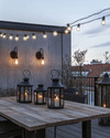 Industrial deck at dusk featuring warm white Edison bulb string lights draped overhead, dark bronze gooseneck wall sconces, a raw wood dining table, and large black metal lanterns with LED candles. — close-up of textures and materials