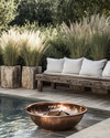 A rustic poolside area featuring a reclaimed timber bench with linen cushions, large natural stone planters filled with tall grasses, and a hammered copper fire bowl, all set against a backdrop of natural stone paving and lush greenery.