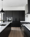 A sleek modern kitchen featuring matte black handleless cabinetry, white quartz countertops, integrated stainless steel appliances, and a minimalist black pendant light over a large kitchen island. — alternate angle