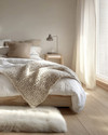 A cozy Scandinavian bedroom featuring a bed dressed in white linen, a chunky knit throw, and a sheepskin rug on light wooden floorboards. Soft, diffused light comes from a simple bedside lamp and a large window.