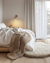 A cozy Scandinavian bedroom featuring a bed dressed in white linen, a chunky knit throw, and a sheepskin rug on light wooden floorboards. Soft, diffused light comes from a simple bedside lamp and a large window. — alternate angle