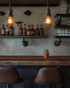 An inviting industrial home bar featuring a reclaimed wood countertop, dark metal pipe shelving, and leather bar stools. Warm Edison bulb pendants cast an amber glow, highlighting vintage amber glass bottles and a small potted succulent. — styling and décor detail