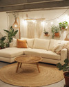 A cozy boho basement living area with a light cream sectional sofa, a round jute rug, and a natural wood coffee table. Warm lighting from a rattan floor lamp and string lights illuminates various potted plants, macrame wall hangings, and terracotta accents.