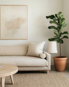 A serene Japandi great room featuring a low-slung oatmeal linen sofa, a light oak round coffee table, and a natural jute rug. Walls are painted in a soft off-white, adorned with a minimalist abstract nature print. A tall Fiddle Leaf Fig in a terracotta pot stands beside a ceramic table lamp with a linen shade, creating a calm and inviting atmosphere. — close-up of textures and materials