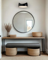 A modern mudroom features a natural oak console table with a minimalist black metal frame, paired with a charcoal grey upholstered bench. Overhead, a matte black sculptural flush-mount light illuminates the space, complemented by a large, frameless circular mirror above the console and woven jute baskets for storage. — styling and décor detail