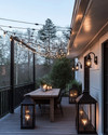 Industrial deck at dusk featuring warm white Edison bulb string lights draped overhead, dark bronze gooseneck wall sconces, a raw wood dining table, and large black metal lanterns with LED candles.