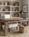A warm farmhouse home office featuring a large distressed oak desk, a cream linen office chair, open shelving with books and woven baskets, and walls painted in a soft greige. A braided jute rug grounds the space, and a matte black task lamp sits on the desk. — styling and décor detail