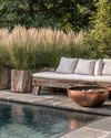 A rustic poolside area featuring a reclaimed timber bench with linen cushions, large natural stone planters filled with tall grasses, and a hammered copper fire bowl, all set against a backdrop of natural stone paving and lush greenery. — styling and décor detail