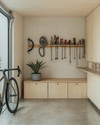 A small Japandi garage featuring light birch plywood cabinetry, a polished concrete floor, and walls painted in a soft off-white. A minimalist bamboo storage bench sits against one wall, with a single snake plant in a grey ceramic pot. Wall-mounted racks hold neatly organized tools, and a bicycle is suspended, maximizing floor space. — styling and décor detail