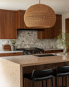 A Mid-Century Modern kitchen featuring flat-panel walnut cabinetry, a honed quartzite island, and a geometric terrazzo backsplash. A large woven rattan pendant light hangs above the island, which is flanked by black leather bar stools, all in soft natural daylight.