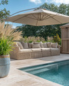 A serene coastal pool area featuring light travertine pavers, deep-seated wicker outdoor sofas with sand-colored cushions, and a large cantilever umbrella providing ample shade. Lush green ornamental grasses in weighty ceramic planters frame the space, while a weathered wood outdoor shower stands nearby. — styling and décor detail