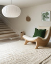 A bright Scandinavian basement living area featuring a light oak sculptural lounge chair, a large cream hand-knotted wool rug, and an oversized white paper lantern pendant light, with subtle forest green textile accents. — styling and décor detail