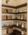 A cozy traditional pantry featuring warm cream walls, rich dark wood shelving, and a mix of woven rattan baskets and clear glass jars holding pantry staples. An aged brass semi-flush mount light fixture illuminates the space. — close-up of textures and materials