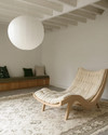 A bright Scandinavian basement living area featuring a light oak sculptural lounge chair, a large cream hand-knotted wool rug, and an oversized white paper lantern pendant light, with subtle forest green textile accents. — alternate angle