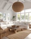 A bright coastal great room featuring a large off-white linen sofa, a reclaimed wood coffee table, and a natural jute area rug. Rattan armchairs flank the space, while a large woven pendant light hangs from the ceiling, all under ample natural light. — alternate angle