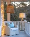 A well-lit coastal porch at dusk, featuring a woven rattan pendant light, string lights draped along the railing, and a floor lamp with a linen shade next to a white wicker chair with soft blue throw pillows. — alternate angle