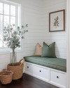 A bright farmhouse mudroom with white shiplap walls, a built-in bench with a sage green cushion, and dark wood flooring. Seasonal touches include a vase of eucalyptus, woven baskets, and a terracotta pot with dried botanicals, all bathed in warm, natural light. — close-up of textures and materials