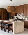 A Mid-Century Modern kitchen featuring flat-panel walnut cabinetry, a honed quartzite island, and a geometric terrazzo backsplash. A large woven rattan pendant light hangs above the island, which is flanked by black leather bar stools, all in soft natural daylight. — close-up of textures and materials