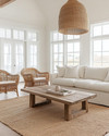 A bright coastal great room featuring a large off-white linen sofa, a reclaimed wood coffee table, and a natural jute area rug. Rattan armchairs flank the space, while a large woven pendant light hangs from the ceiling, all under ample natural light. — styling and décor detail