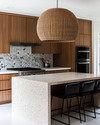 A Mid-Century Modern kitchen featuring flat-panel walnut cabinetry, a honed quartzite island, and a geometric terrazzo backsplash. A large woven rattan pendant light hangs above the island, which is flanked by black leather bar stools, all in soft natural daylight. — styling and décor detail