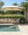 A serene coastal pool area featuring light travertine pavers, deep-seated wicker outdoor sofas with sand-colored cushions, and a large cantilever umbrella providing ample shade. Lush green ornamental grasses in weighty ceramic planters frame the space, while a weathered wood outdoor shower stands nearby. — close-up of textures and materials